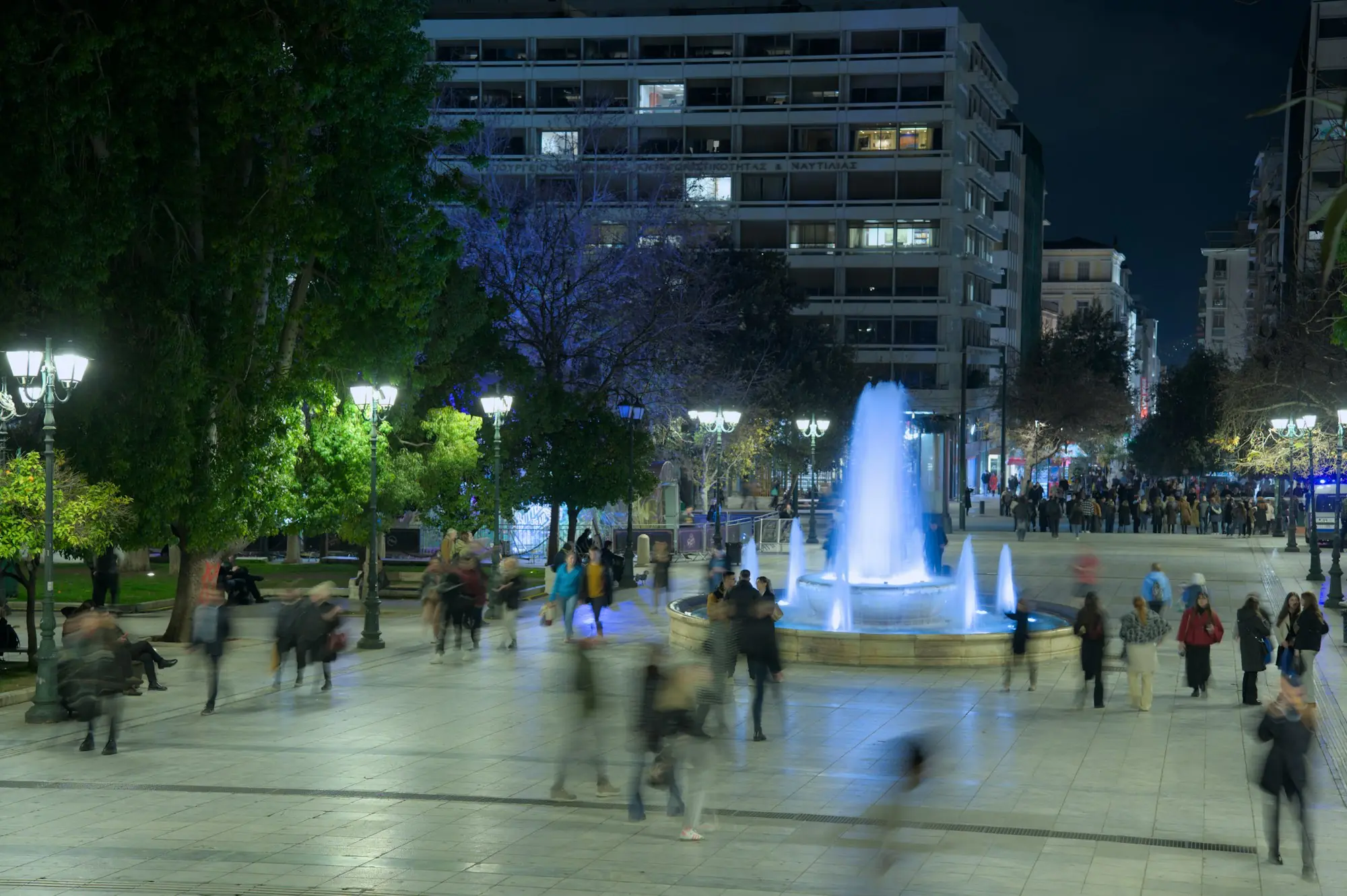 People crossing a lit city square with a fountain at night